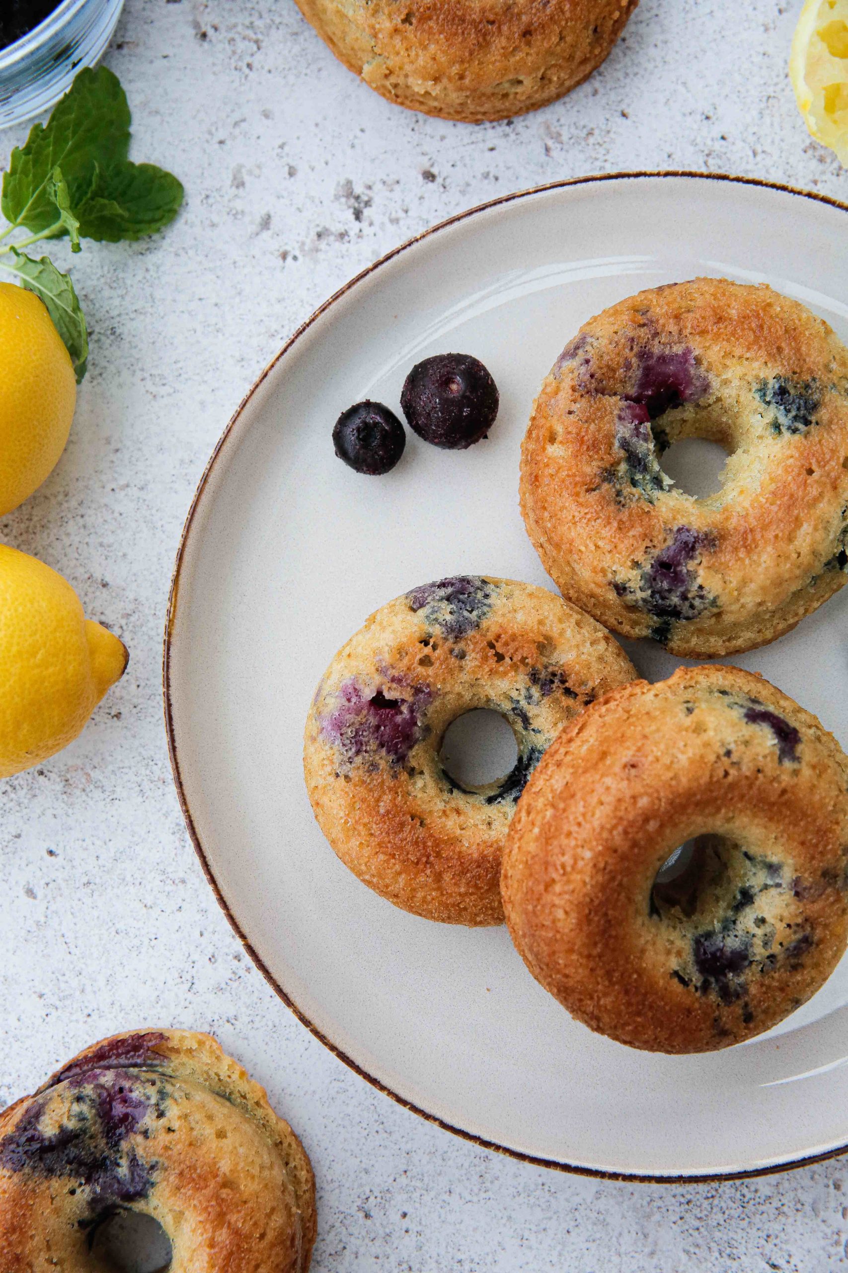 These baked donuts, flavoured with lemon and studded with juicy blueberries are squidgy perfection! They're non yeast, more cake donuts which means they're super simple and quick to make. Absolutely delicious warm or cold with a cuppa for breakfast or an afternoon vegan treat! Recipe on thecookandhim.com #vegandonut #bakeddonuts #homemadedonuts #cakedonuts