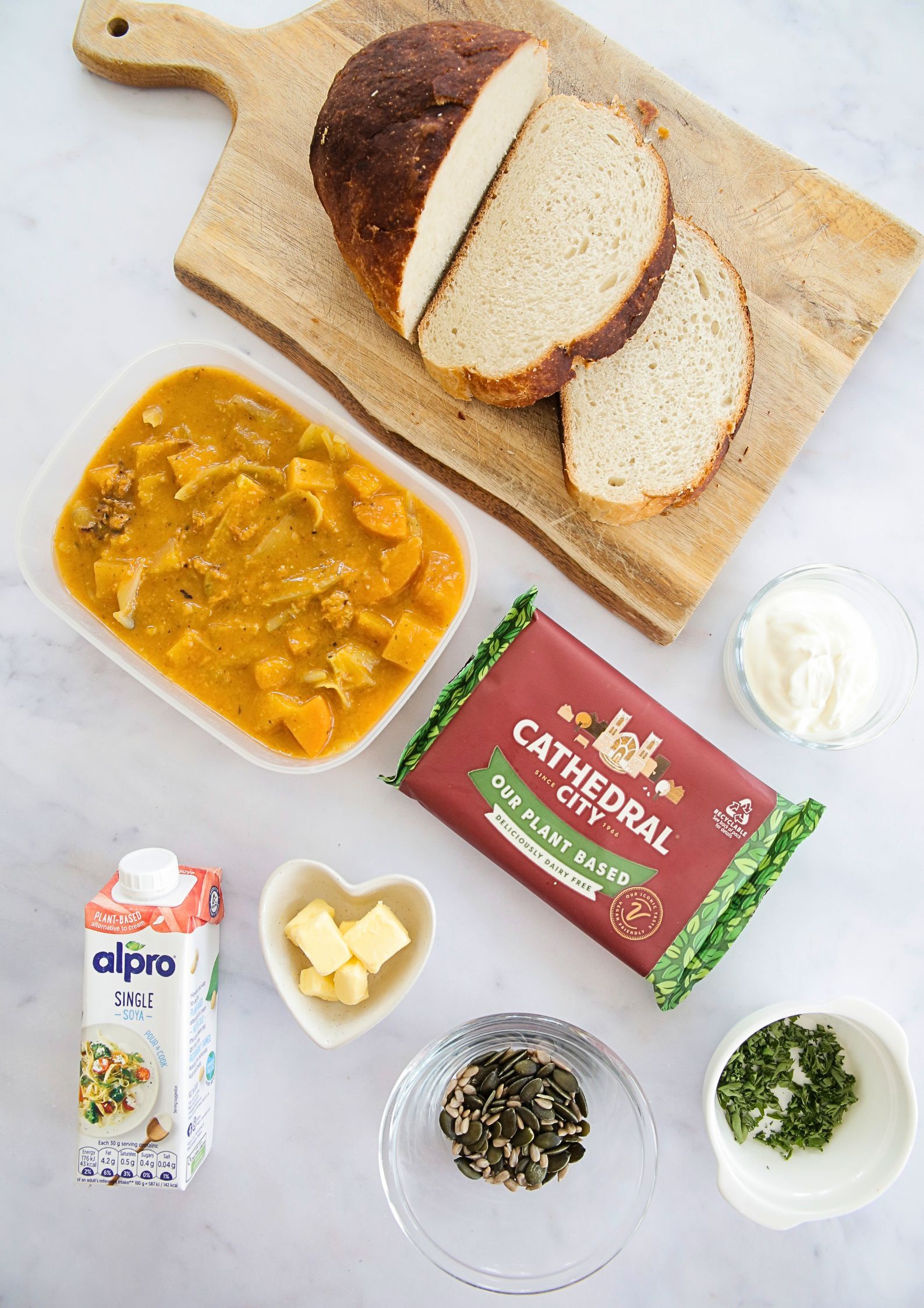 Top down photo of ingredients for soup and a sandwich - a loaf of bread partially sliced on a wooden board, a tub of chunky soup, a wrapped block of vegan cheese, a carton of vegan cream, a bowl of yoghurt, a bowl of diced butter, a dish of fresh herbs and a bowl of toasted seeds