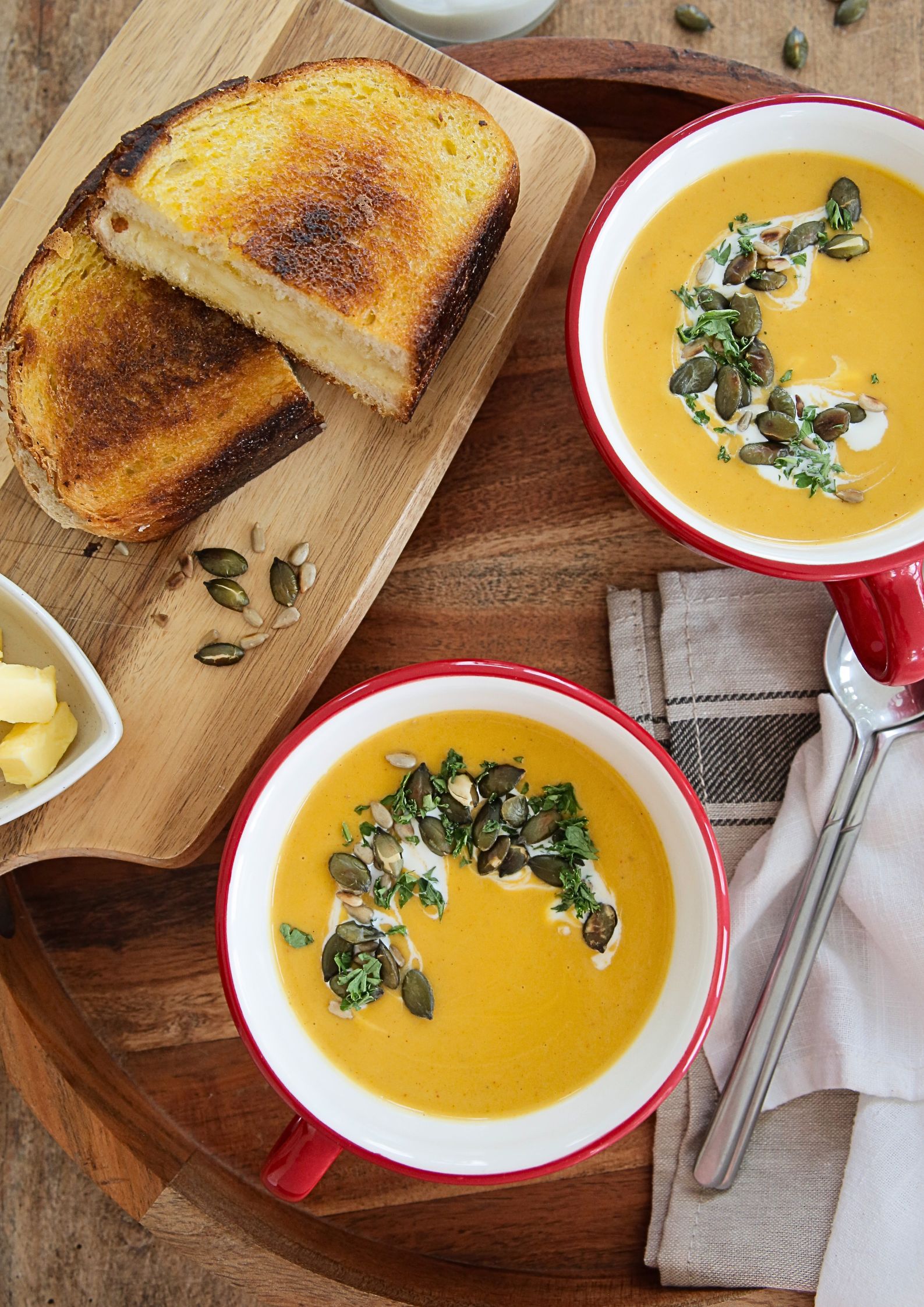 A top down photo of 2 bowls of prepared vegetable soup, yellow in colour and garnished with a yoghurt swirl, toasted seeds and chopped fresh herbs. To the side is a toasted cheese sandwich