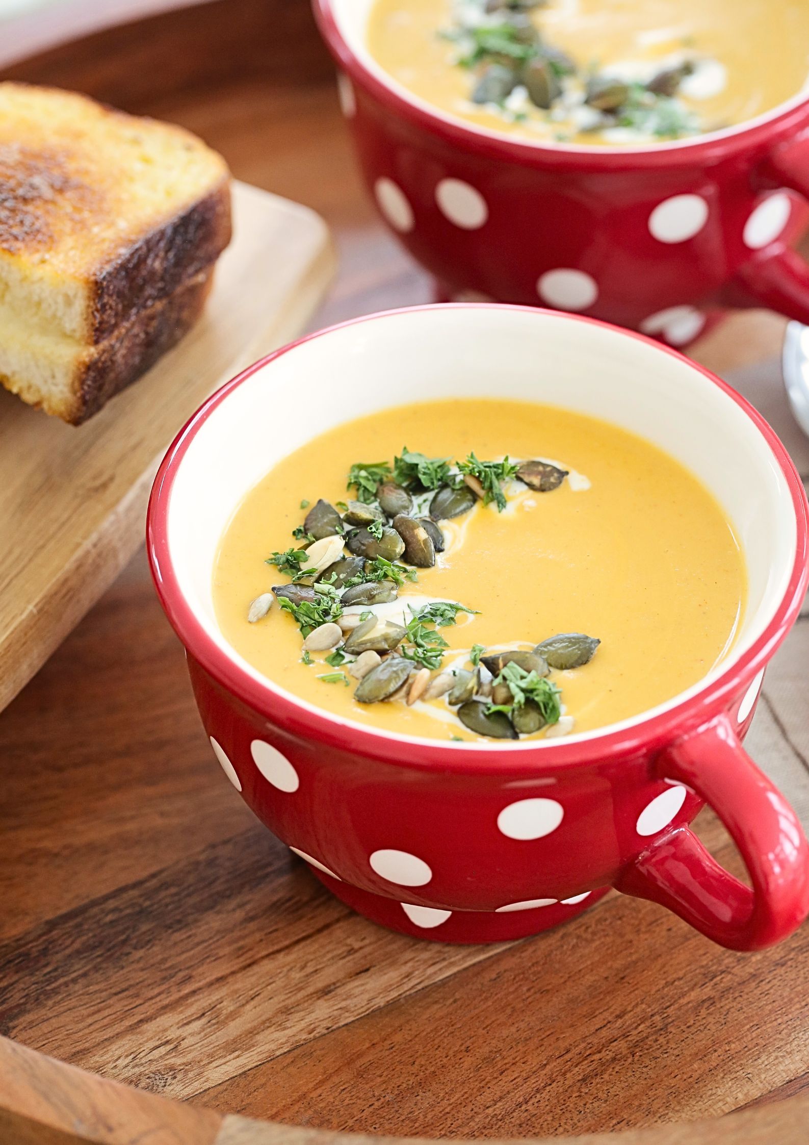 A top down photo of 2 bowls of prepared vegetable soup, yellow in colour and garnished with a yoghurt swirl, toasted seeds and chopped fresh herbs. To the side is a toasted cheese sandwich