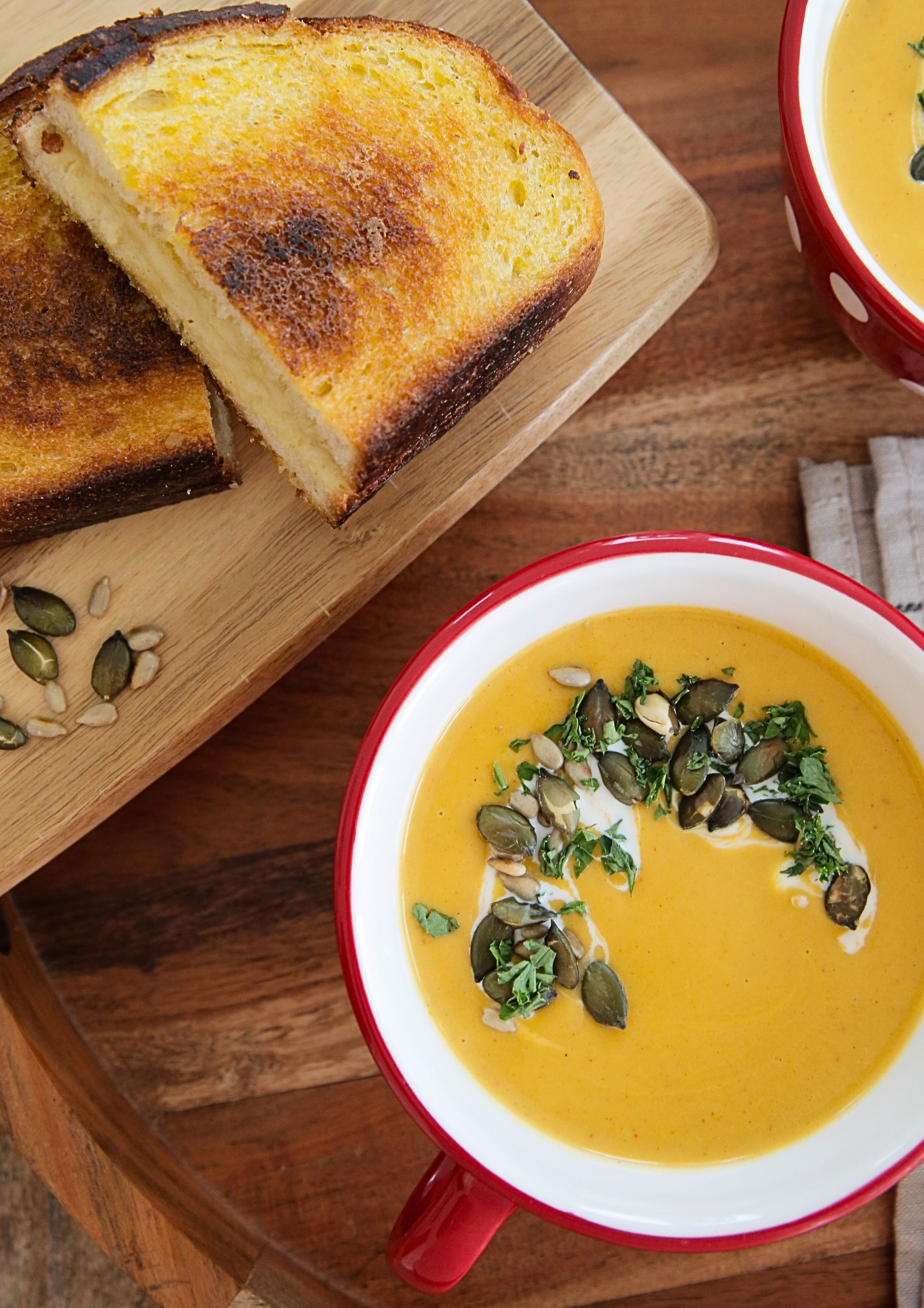 A top down photo of 2 bowls of prepared vegetable soup, yellow in colour and garnished with a yoghurt swirl, toasted seeds and chopped fresh herbs. To the side is a toasted cheese sandwich