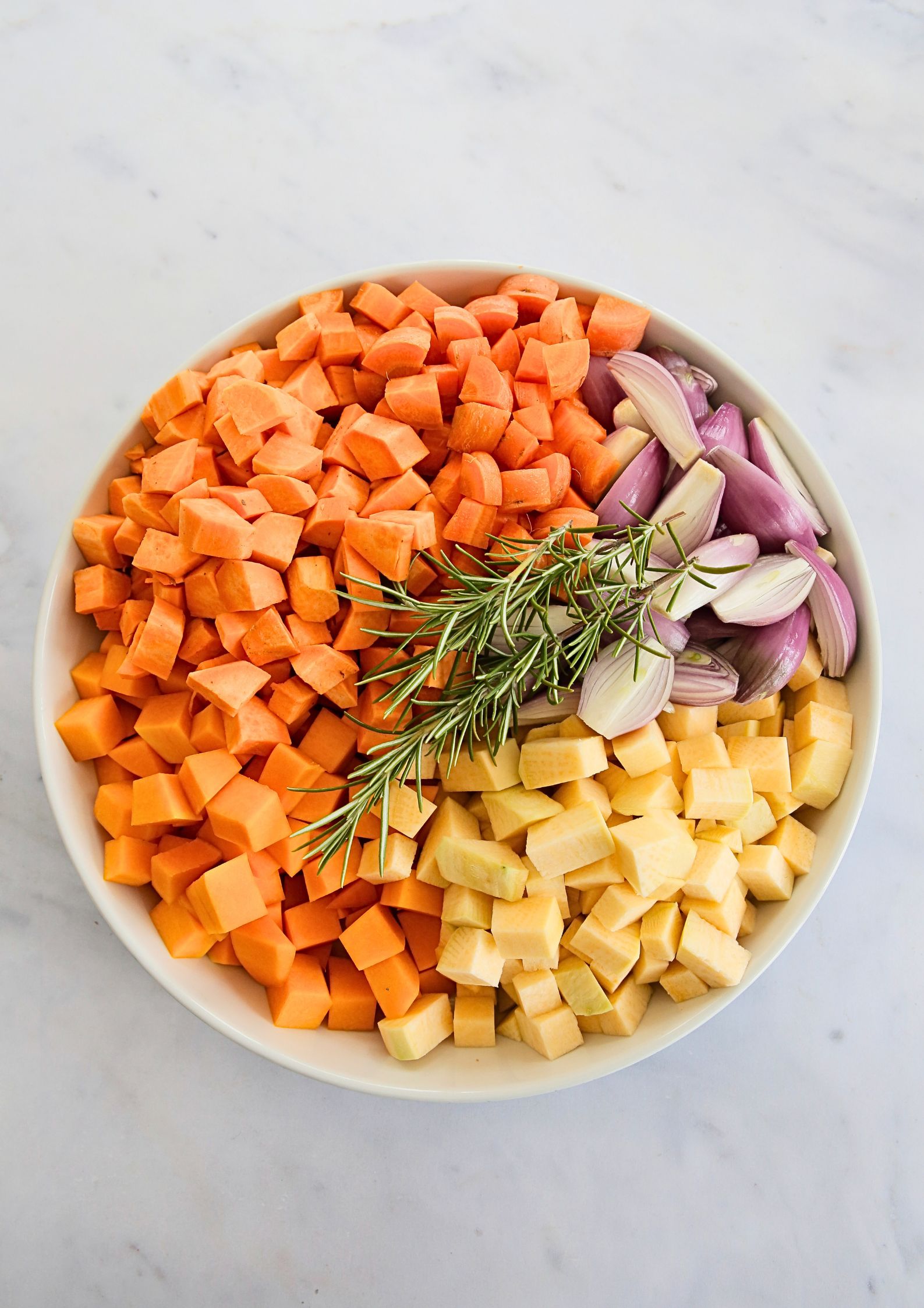 A top down photo of prepared raw vegetables including diced, peeled carrot, butternut squash, sweet potatoes and swede and peeled and halved banana shallots. Lying across the top are sprigs of fresh rosemary. The vegetables are arranged according to colour