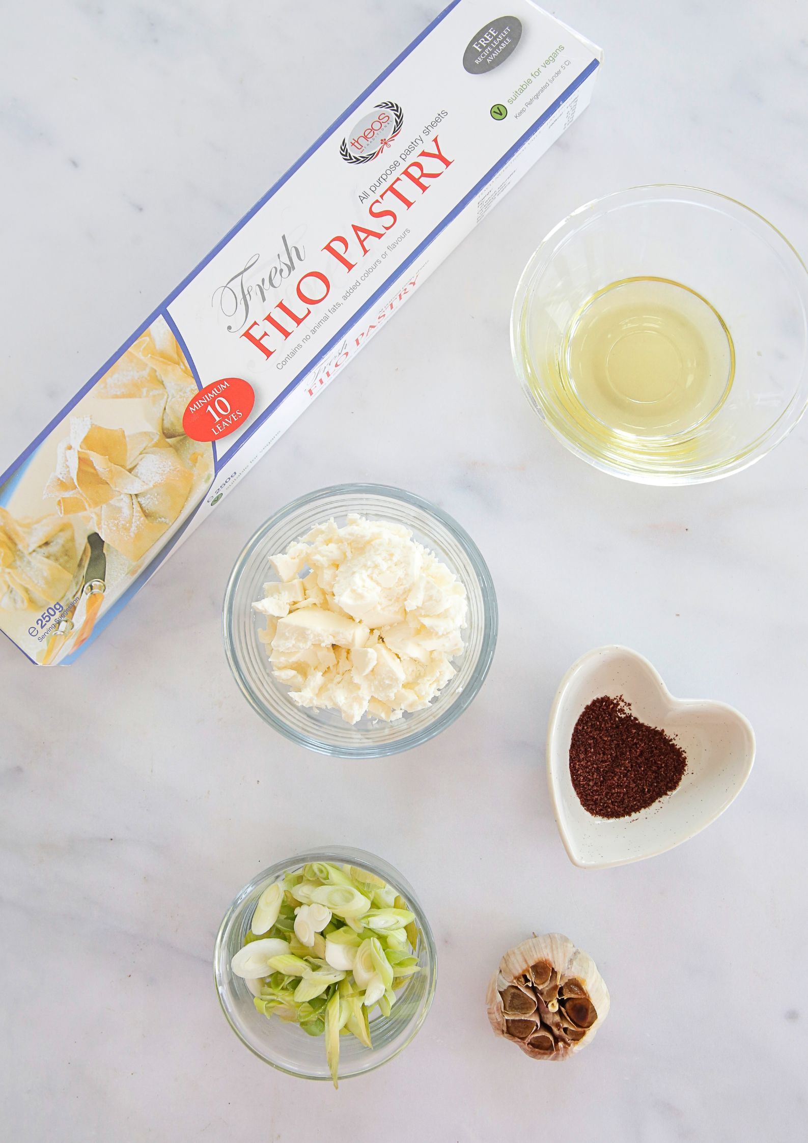 Flat-lay of ingredients to make the feta filo parcels - a pack of filo pastry, a bulb of garlic and bowls of crumbled feta, oil, spices and sliced spring onions