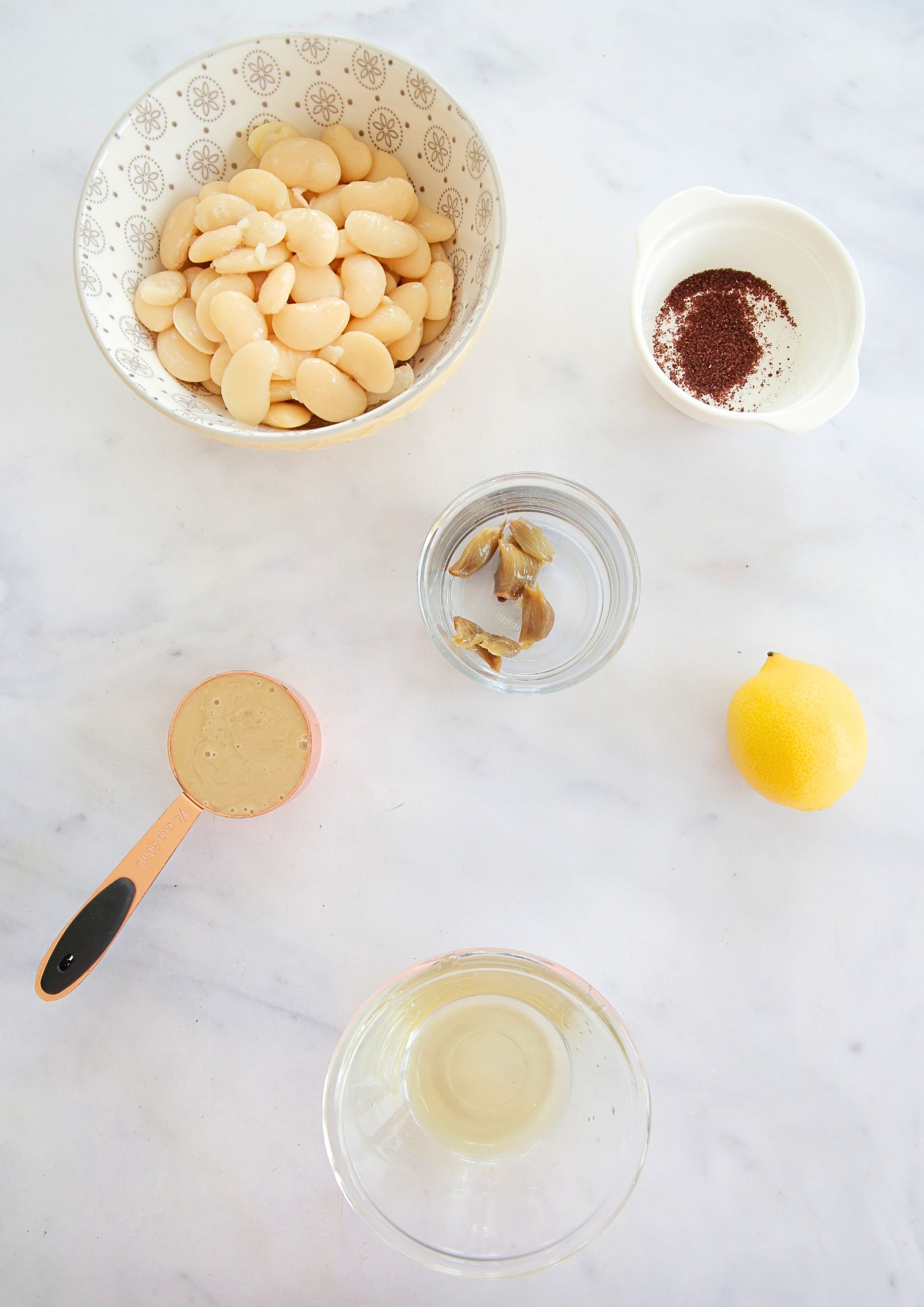 Flat lay of ingredients to make a white bean dip, including a whole lemon and bowls of butterbeans, spices, roast garlic, oil and tahini