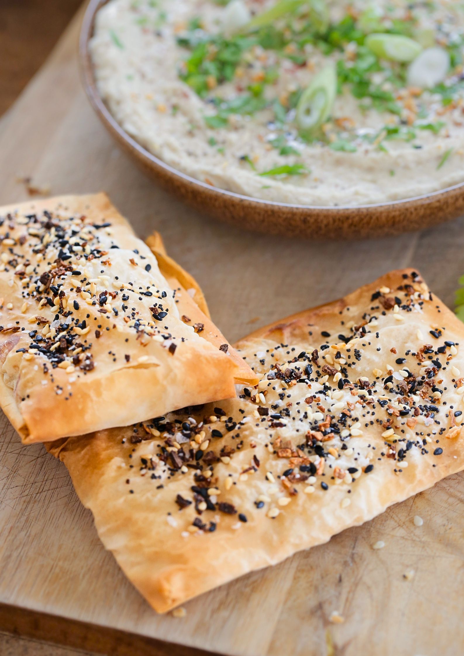 A close up of white bean dip dressed with herbs, spices and spring onions and 2 crispy filo parcels coated in mixed seeds