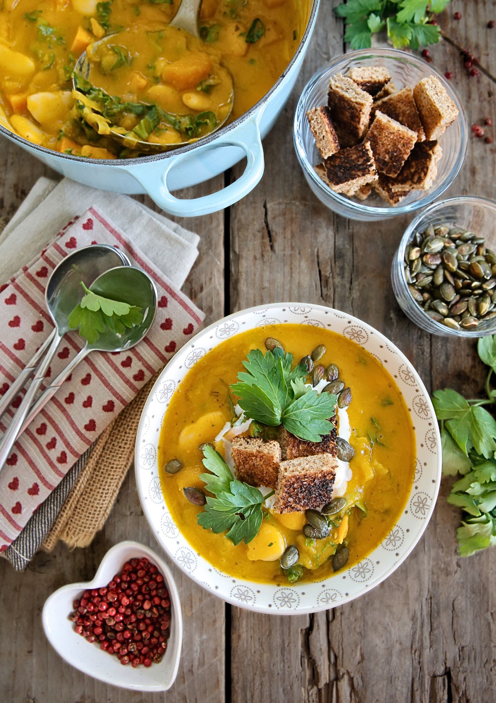 Top down photo of a bowl of autumn vegetable and white bean soup garnished with crusty croutons and fresh herbs. There's also smaller bowls of croutons, pumpkin seeds and peppercorns as well as a saucepan of the soup. Two spoons sit atop a small pile of napkins