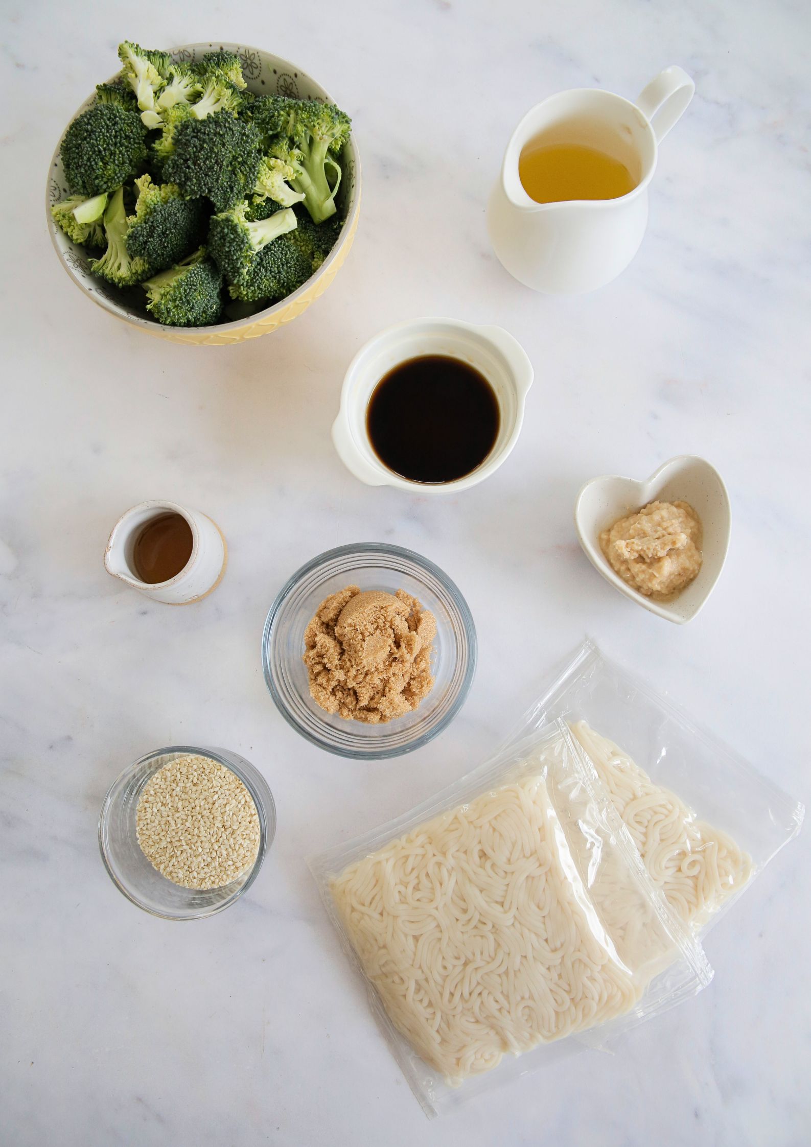 Flat lay of ingredients in bowls to make vegan teriyaki noodles including raw broccoli florets, soy sauce and brandy, sesame oil, brown sugar, sesame seeds and minced ginger and garlic. There's also a jug of pineapple juice and two packets of raw noodles