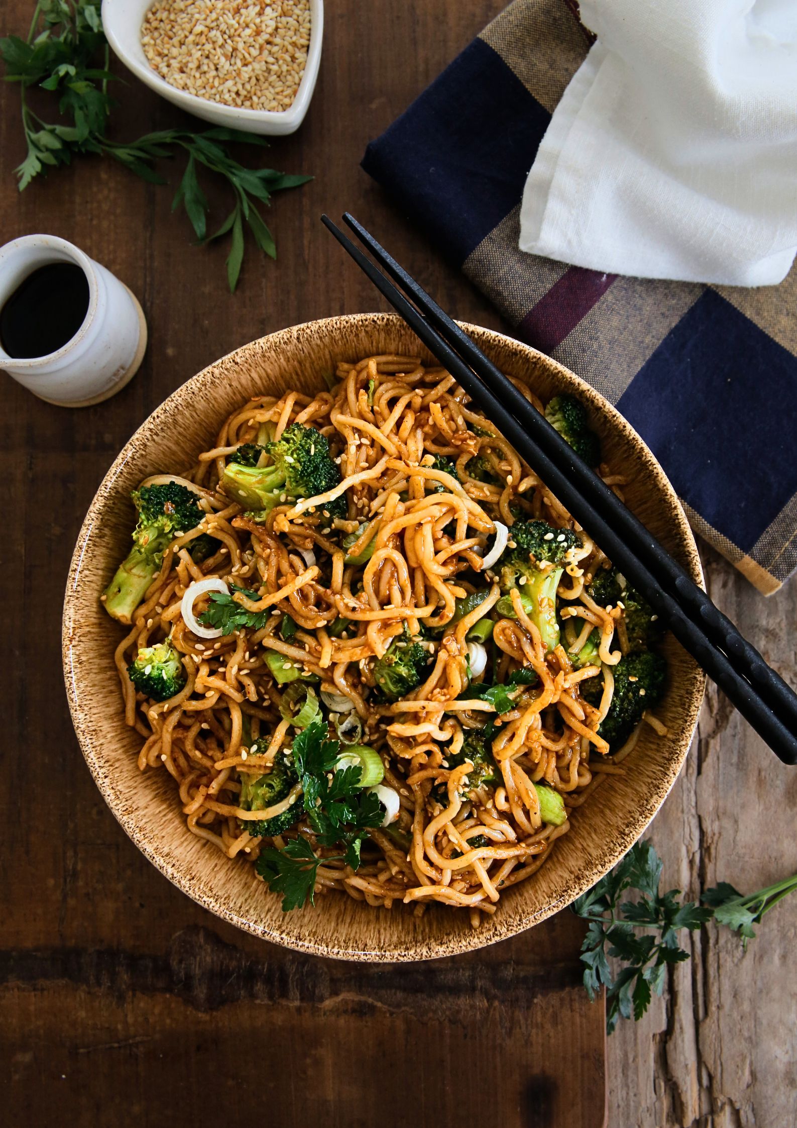 A top down photo of cooked teriyaki noodles in a bowl with chopsticks. A small jug of soy sauce and a dish of toasted sesame seeds are alongside the bowl