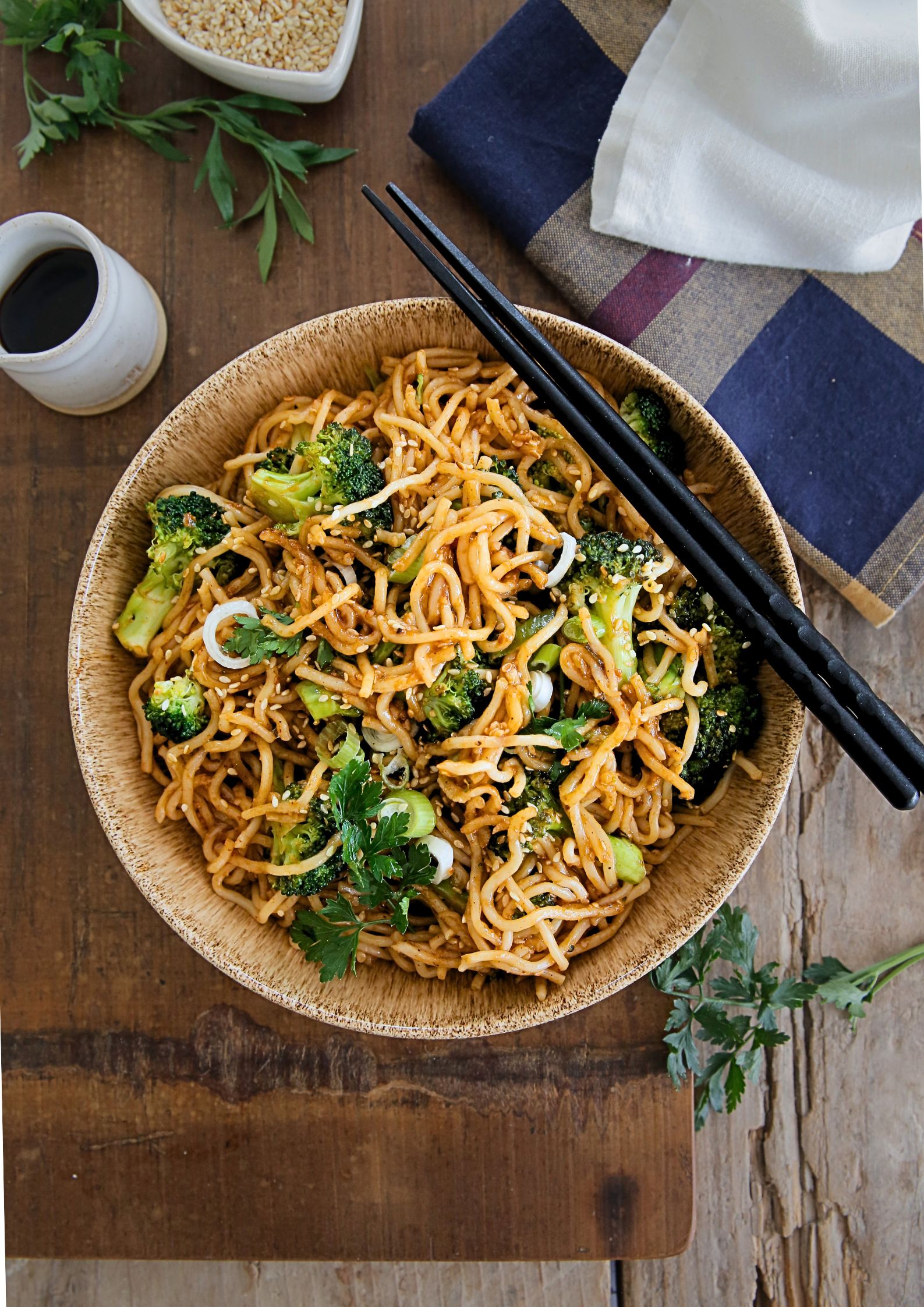 A top down photo of cooked teriyaki noodles in a bowl with chopsticks. A small jug of soy sauce and a dish of toasted sesame seeds are alongside the bowl