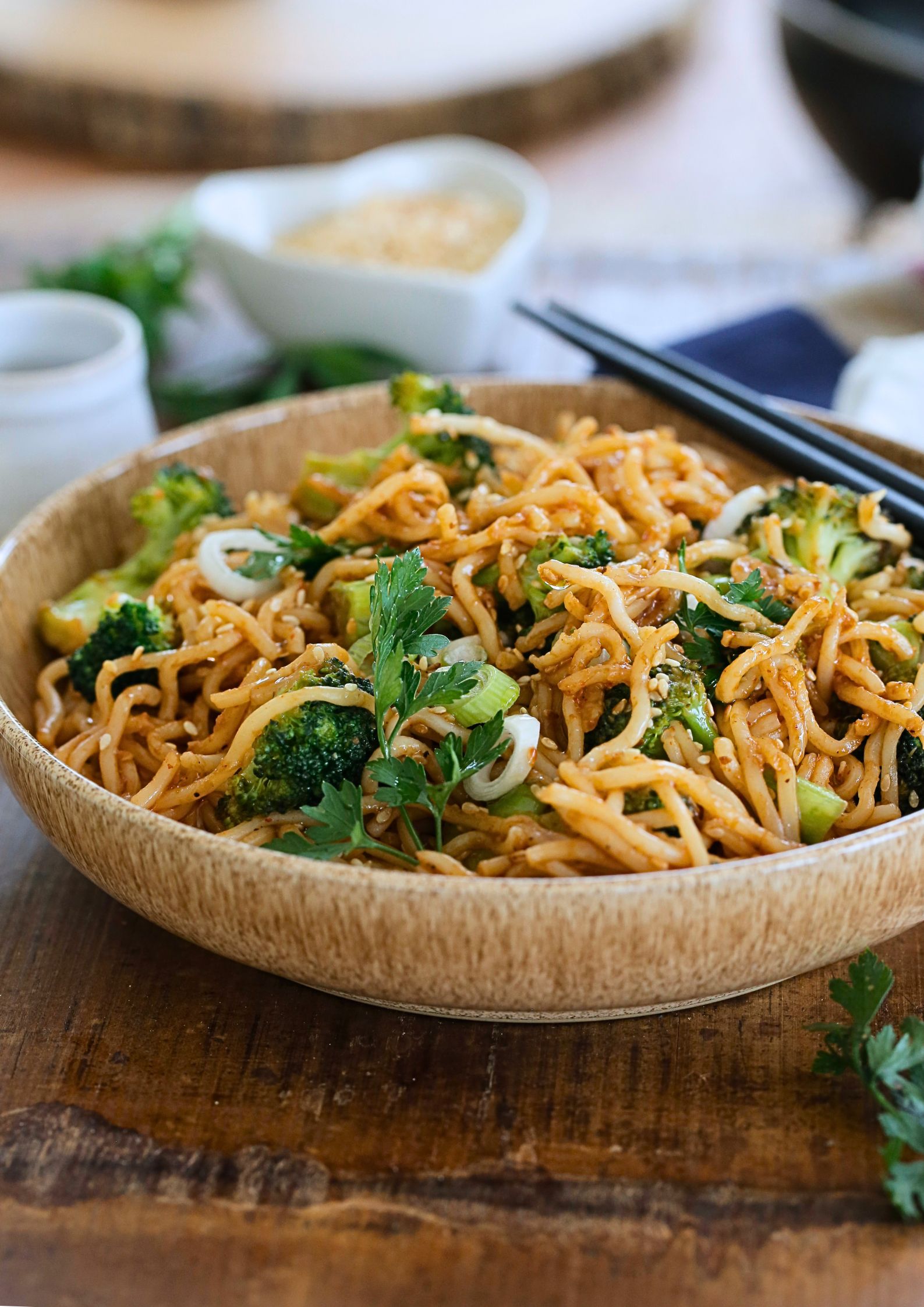 A photo of cooked teriyaki noodles in a bowl with chopsticks. A small jug of soy sauce and a dish of toasted sesame seeds are alongside the bowl