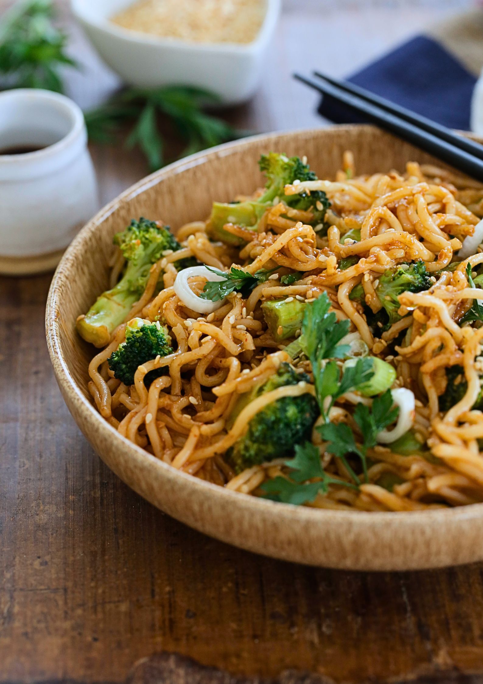 A photo of cooked teriyaki noodles in a bowl with chopsticks. A small jug of soy sauce and a dish of toasted sesame seeds are alongside the bowl