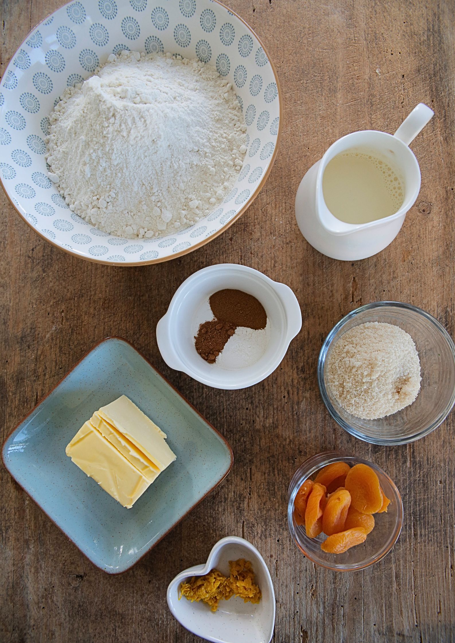 Top down photo of ingredients on a wooden background. There is a bowl of flour, a jug of milk, a small bowl of spice, a bowl of sugar, a small bowl of grated orange zest and a bowl of dried whole apricots. There's also a square plate with some vegan butter