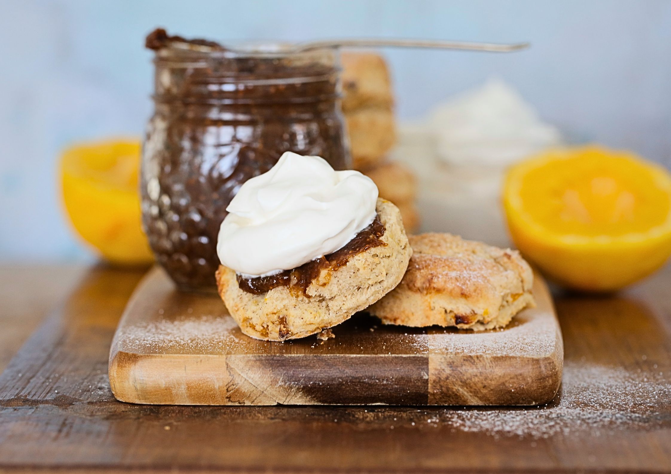 Close up photo of a scone, split and spread with fig jam and topped with whipped vegan cream. The jar of jam and an orange is in the background