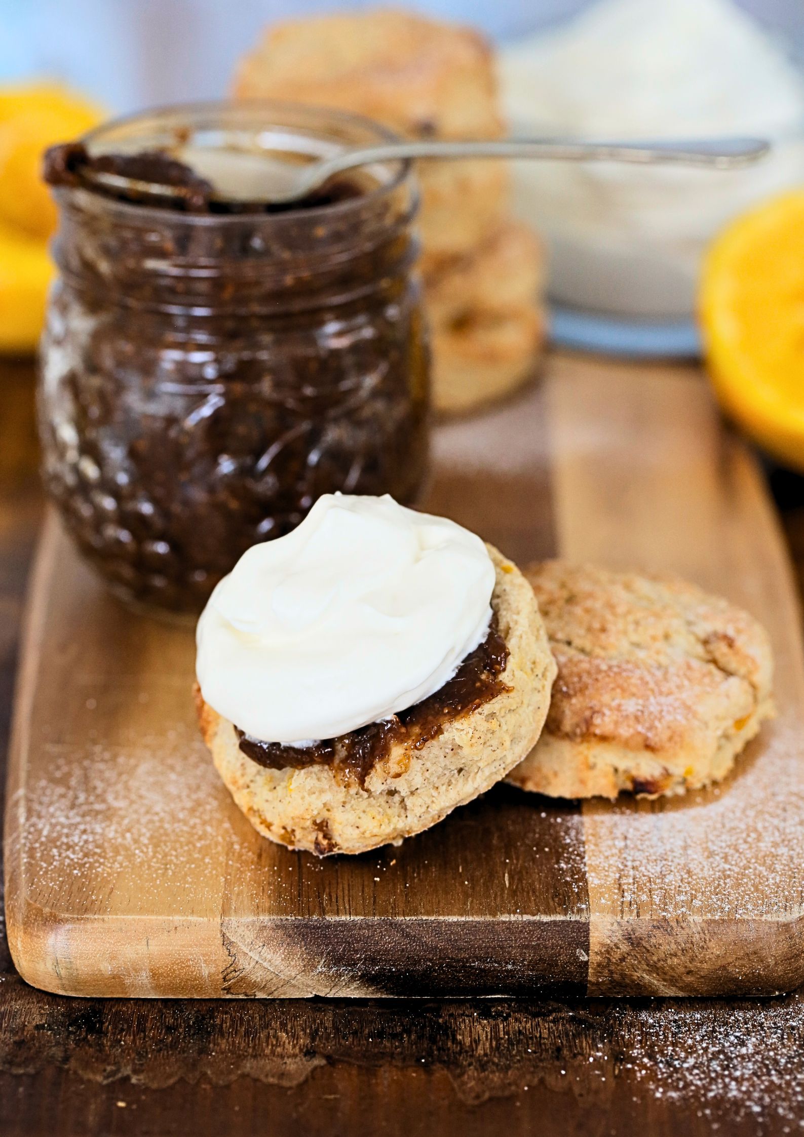 Close up photo of a scone, split and spread with fig jam and topped with whipped vegan cream. The jar of jam and an orange is in the background