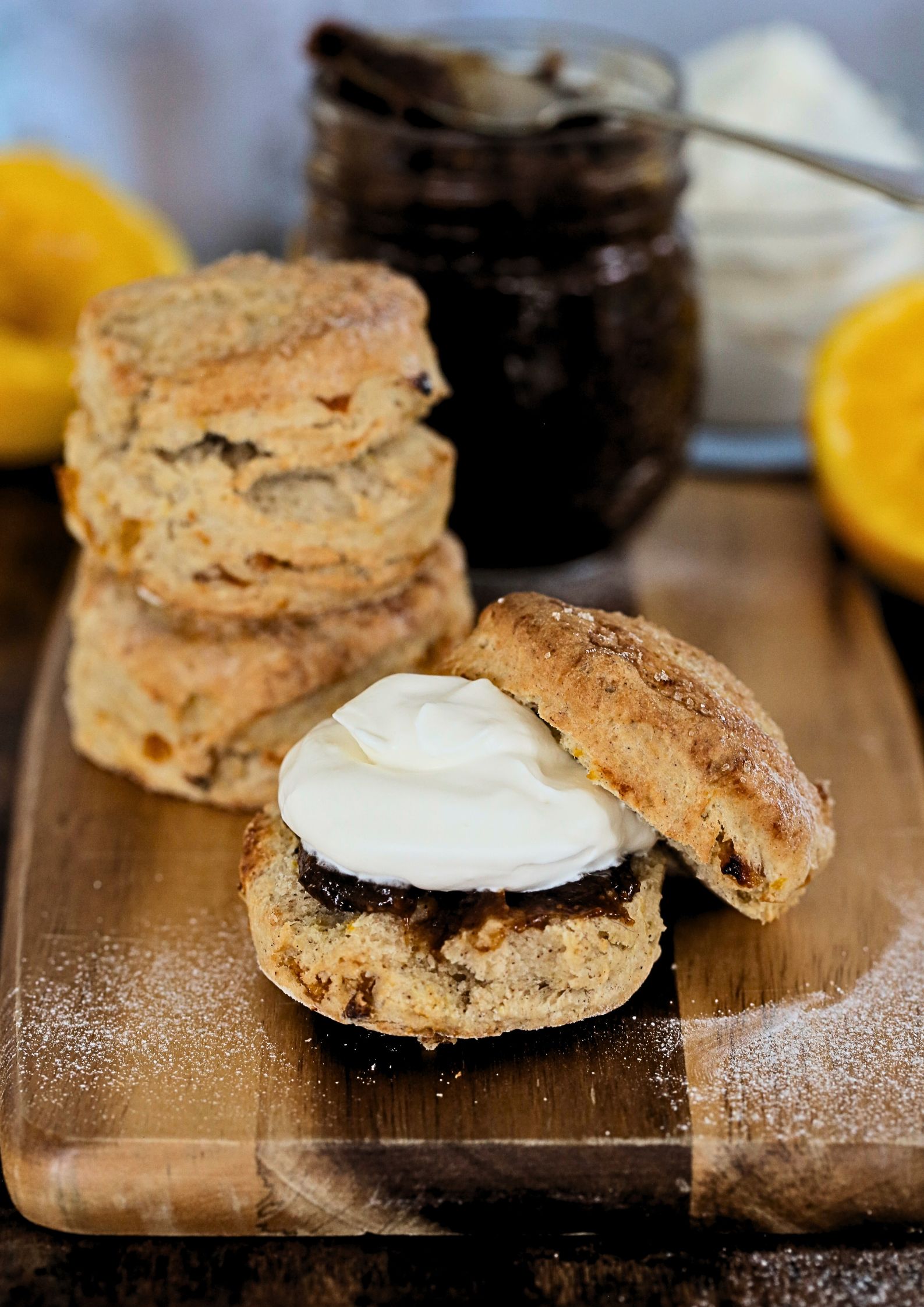 Close up photo of a scone, split and spread with fig jam and topped with whipped vegan cream. The jar of jam and an orange is in the background