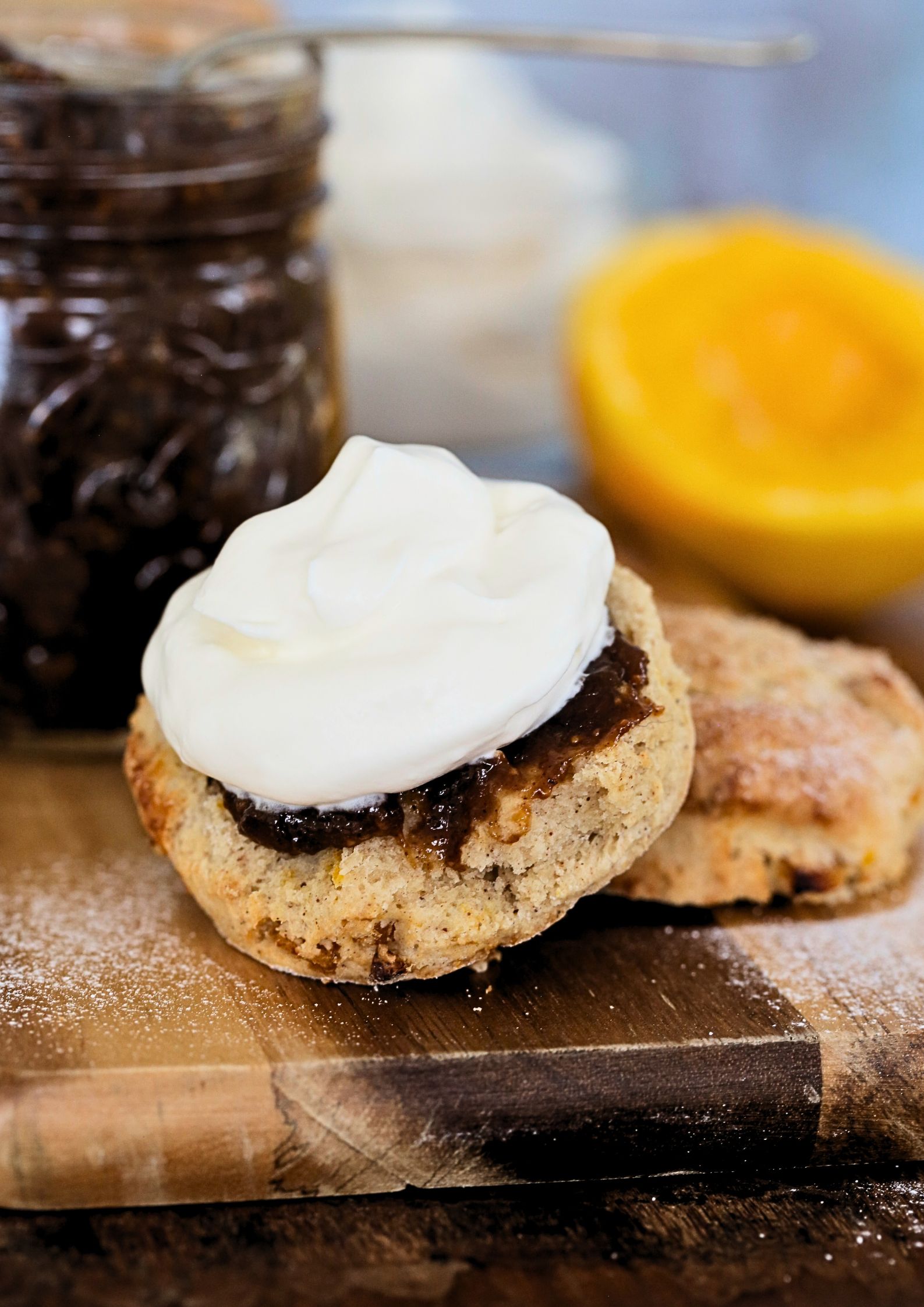 Close up photo of a scone, split and spread with fig jam and topped with whipped vegan cream. The jar of jam and an orange is in the background