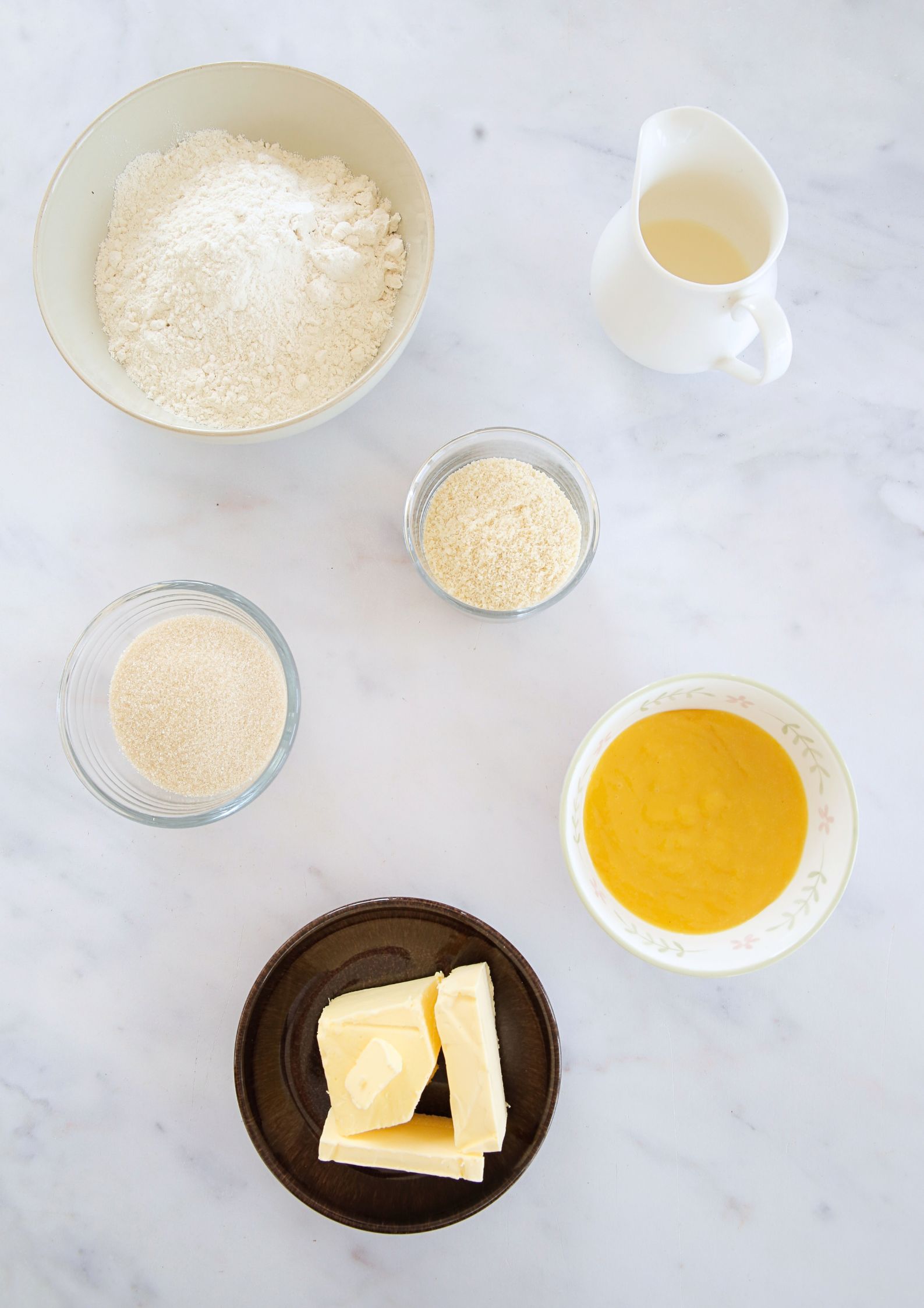 Flat lay on a marble background of various rustic bowls containing flour, caster sugar, ground almonds and vegan butter. There is also a bowl of vegan lemon curd and a small jug of milk