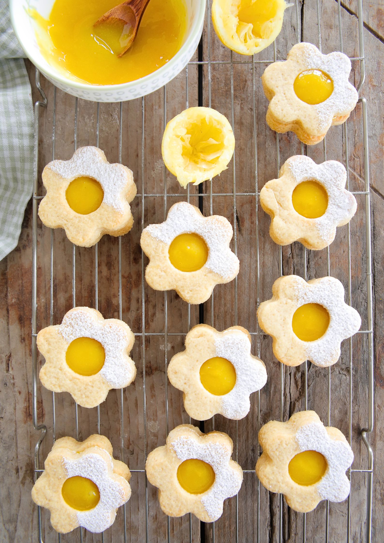 Vegan daisy-shaped shortbread cookies filled with bright yellow lemon curd, dusted with powdered sugar, cooling on a wire rack. A bowl of vegan lemon curd and squeezed lemons sit nearby on a rustic wooden surface.