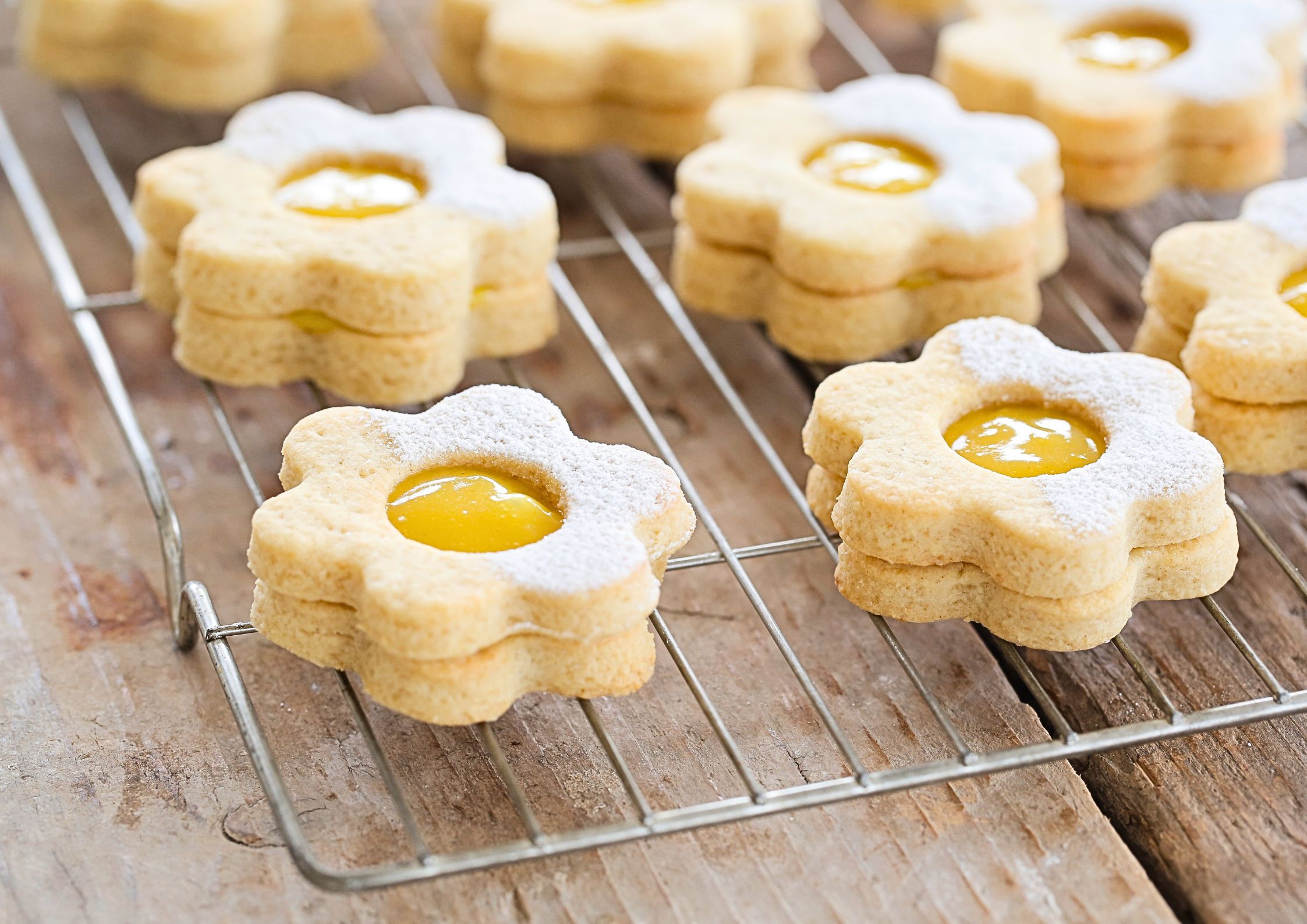 Vegan daisy-shaped shortbread cookies filled with bright yellow lemon curd, dusted with powdered sugar, cooling on a wire rack.