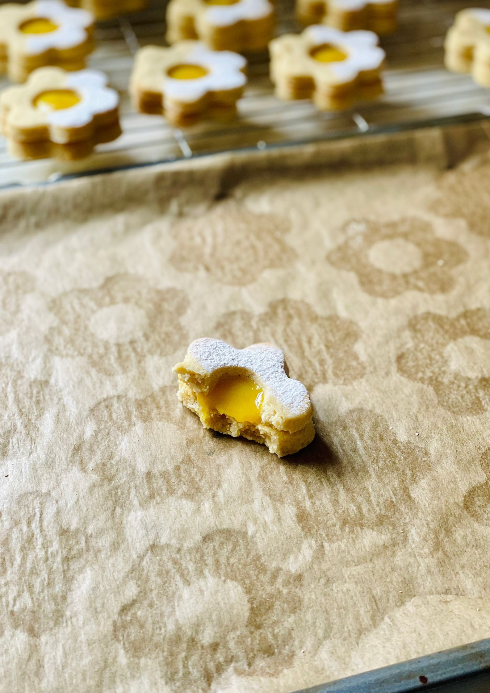 Baked flower-shaped vegan cookies filled with lemon curd, one bitten into, resting on parchment paper with whole cookies on a wire rack in the background