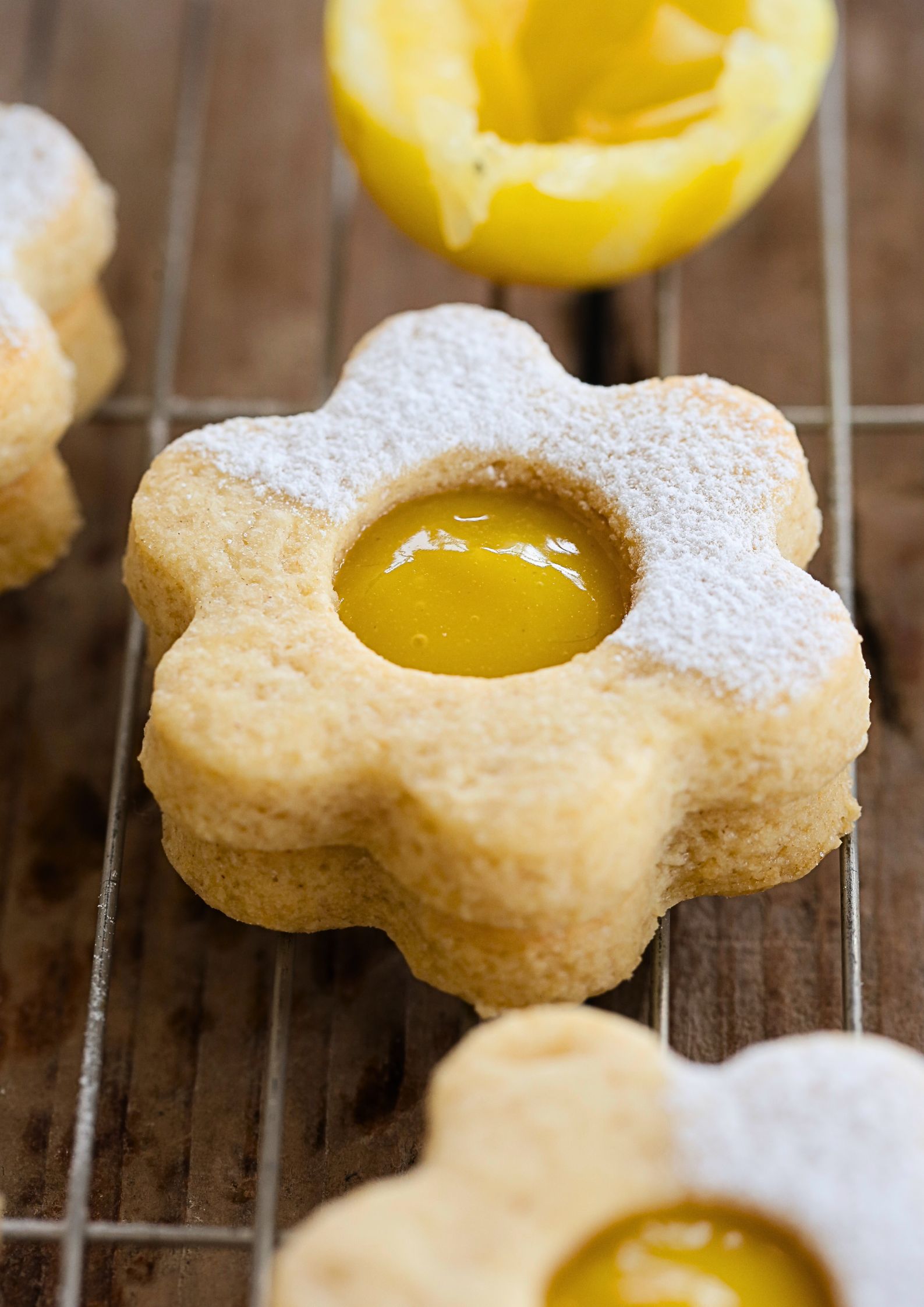 Close up of vegan daisy-shaped shortbread cookies filled with bright yellow lemon curd, dusted with powdered sugar, cooling on a wire rack.