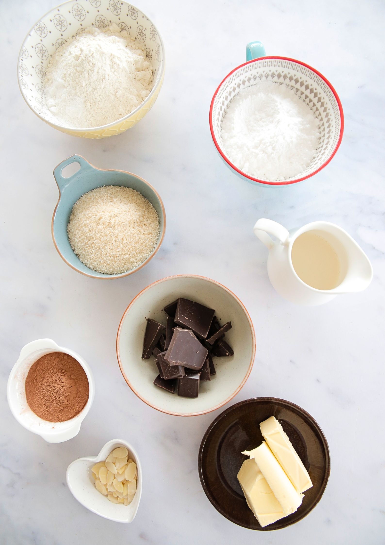 A flat lay of vegan baking ingredients on a marble surface, including flour, sugar, cocoa powder, dark chocolate chunks, vegan butter, plant-based milk, and sliced almonds.