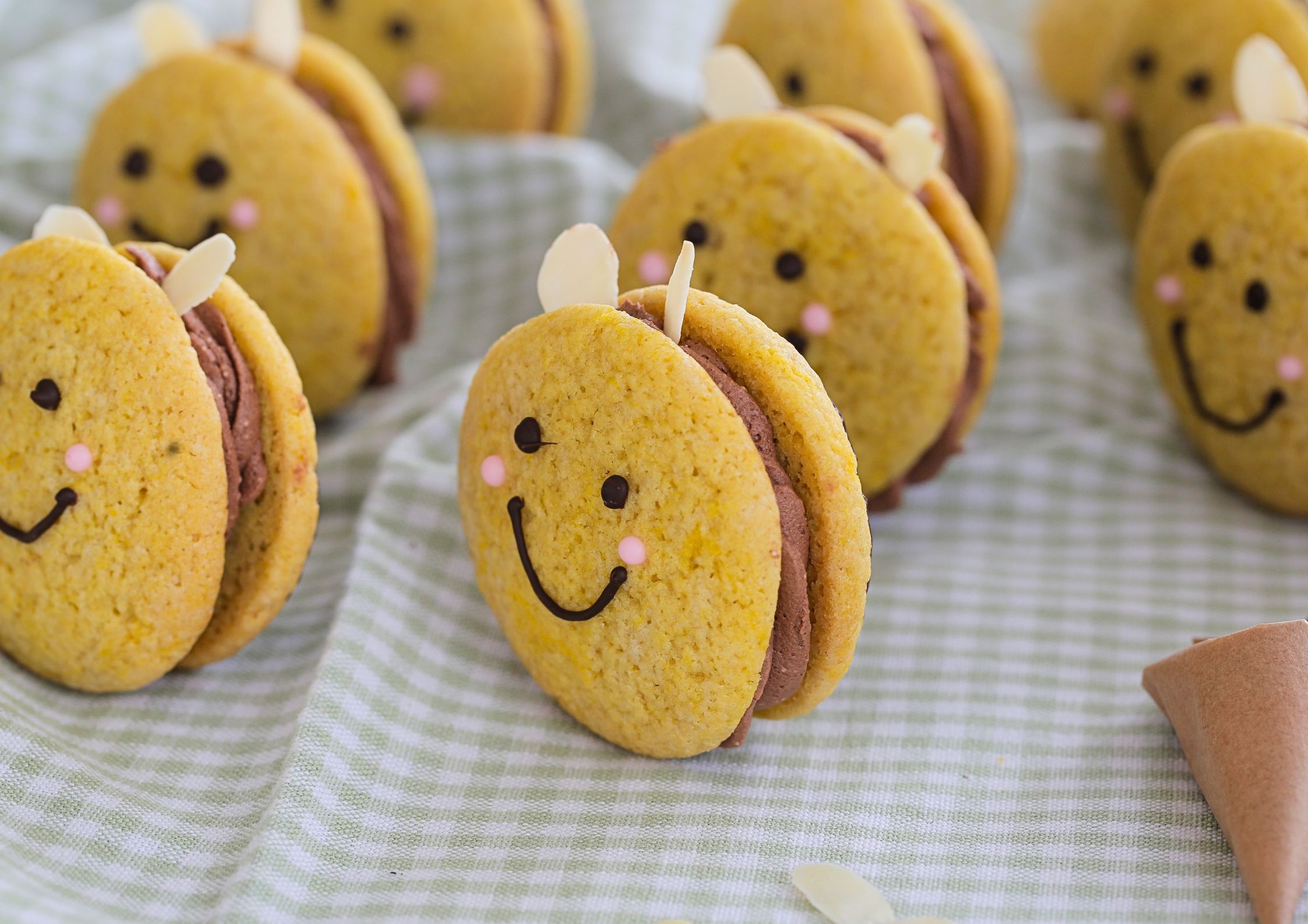 A close-up of an adorable vegan bee cookie with a chocolate smiley face, pink cheeks, and almond slice wings, sandwiched with chocolate buttercream. Other bee cookies are slightly blurred in the background on a green gingham cloth.