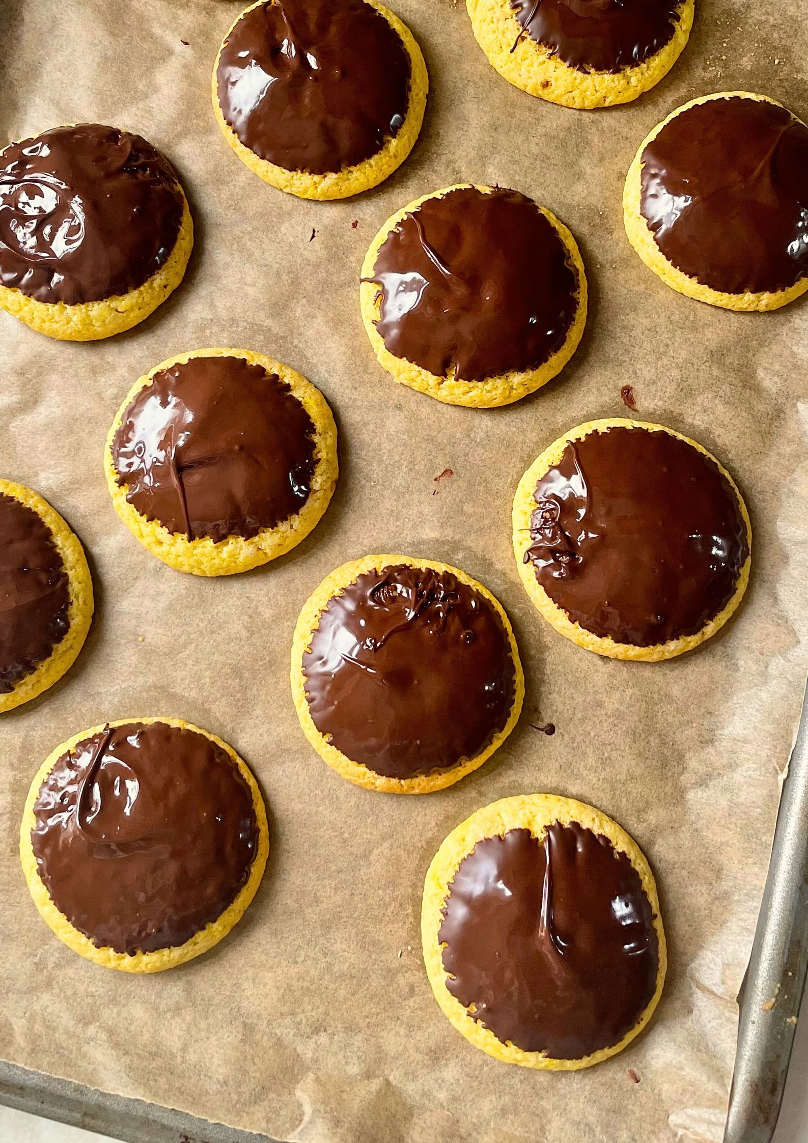 Flat lay of baked yellow cookies just dipped in glistening melted chocolate on a baking tray