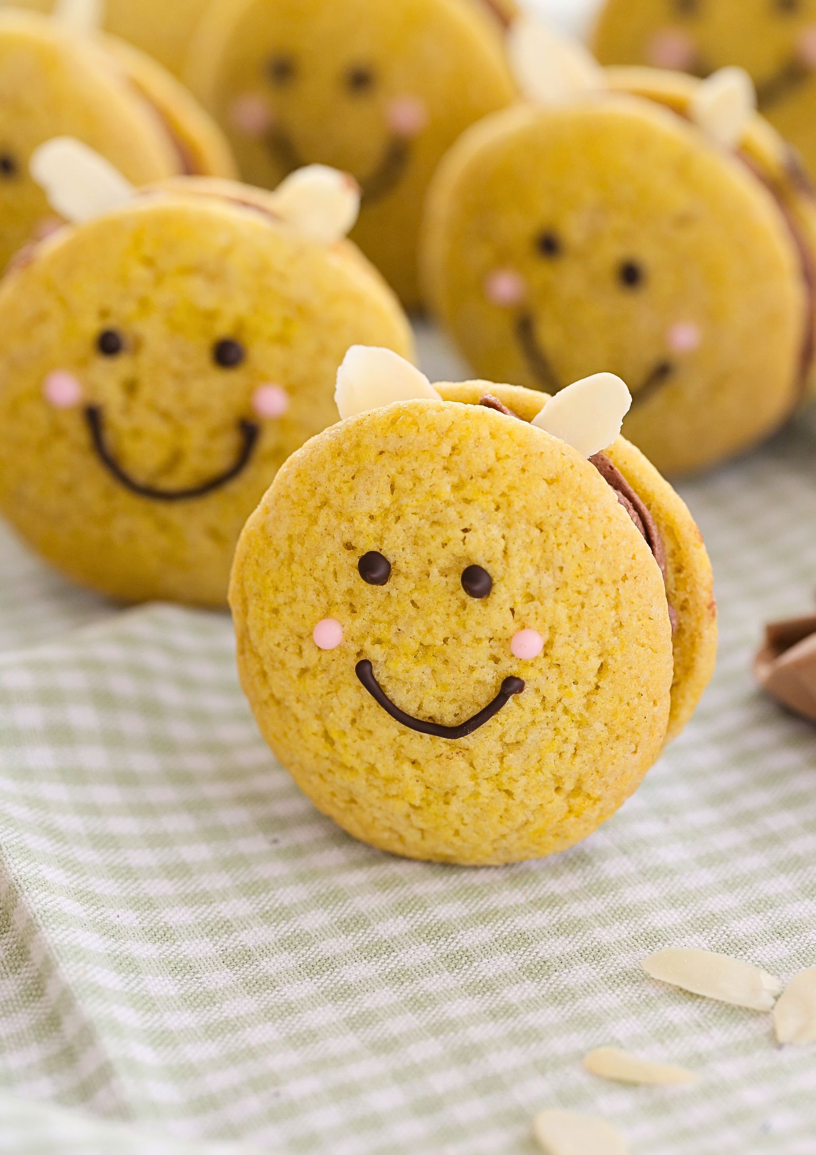 A close-up of an adorable vegan bee cookie with a chocolate smiley face, pink cheeks, and almond slice wings, sandwiched with chocolate buttercream. Other bee cookies are slightly blurred in the background on a green gingham cloth.