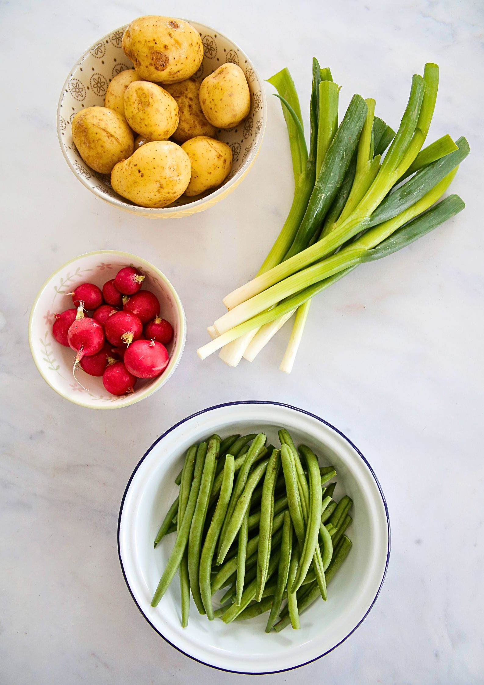 Flat lay on a marble background of various rustic bowls containing raw new potatoes, trimmed green beans and radishes. A bunch of trimmed spring onions sits next to the bowls.