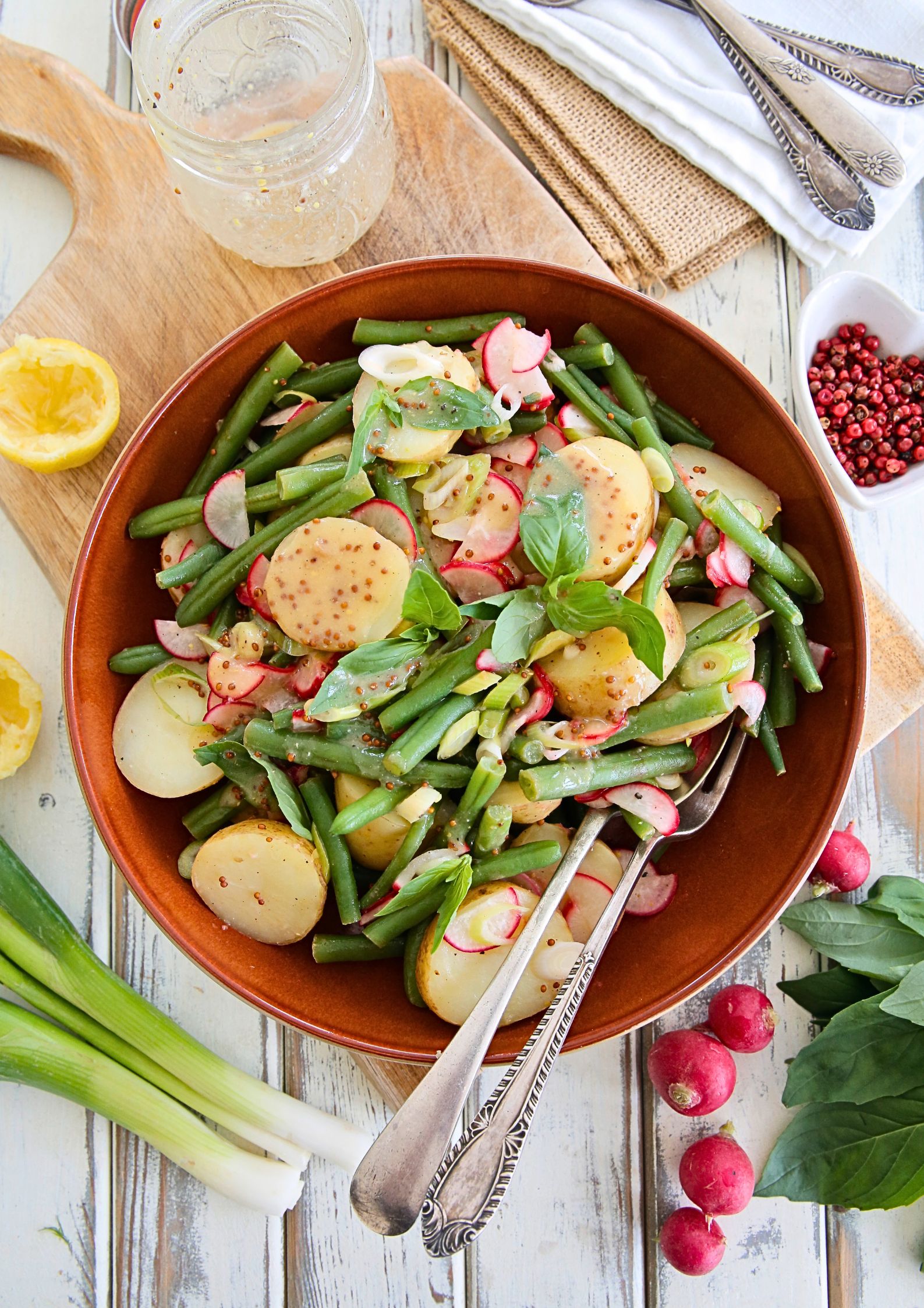 Warm spring salad in a brown bowl featuring boiled new potatoes, green beans, sliced radishes, and fresh basil, drizzled with a miso lemon dressing. Surrounded by fresh ingredients like radishes, spring onions, and lemon on a rustic wooden surface.