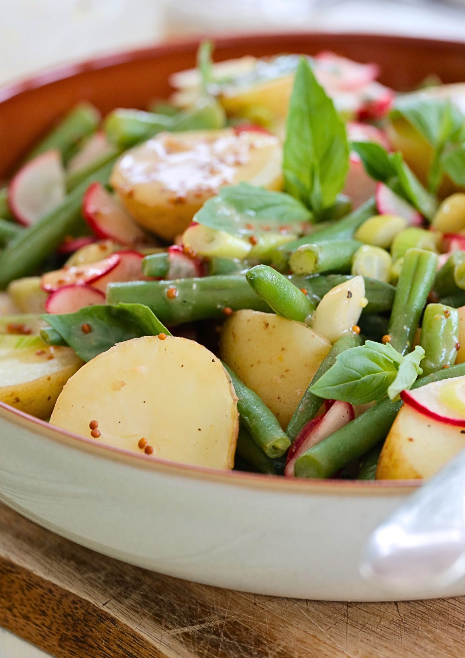 Warm spring salad in a brown bowl featuring boiled new potatoes, green beans, sliced radishes, and fresh basil, drizzled with a miso lemon dressing.