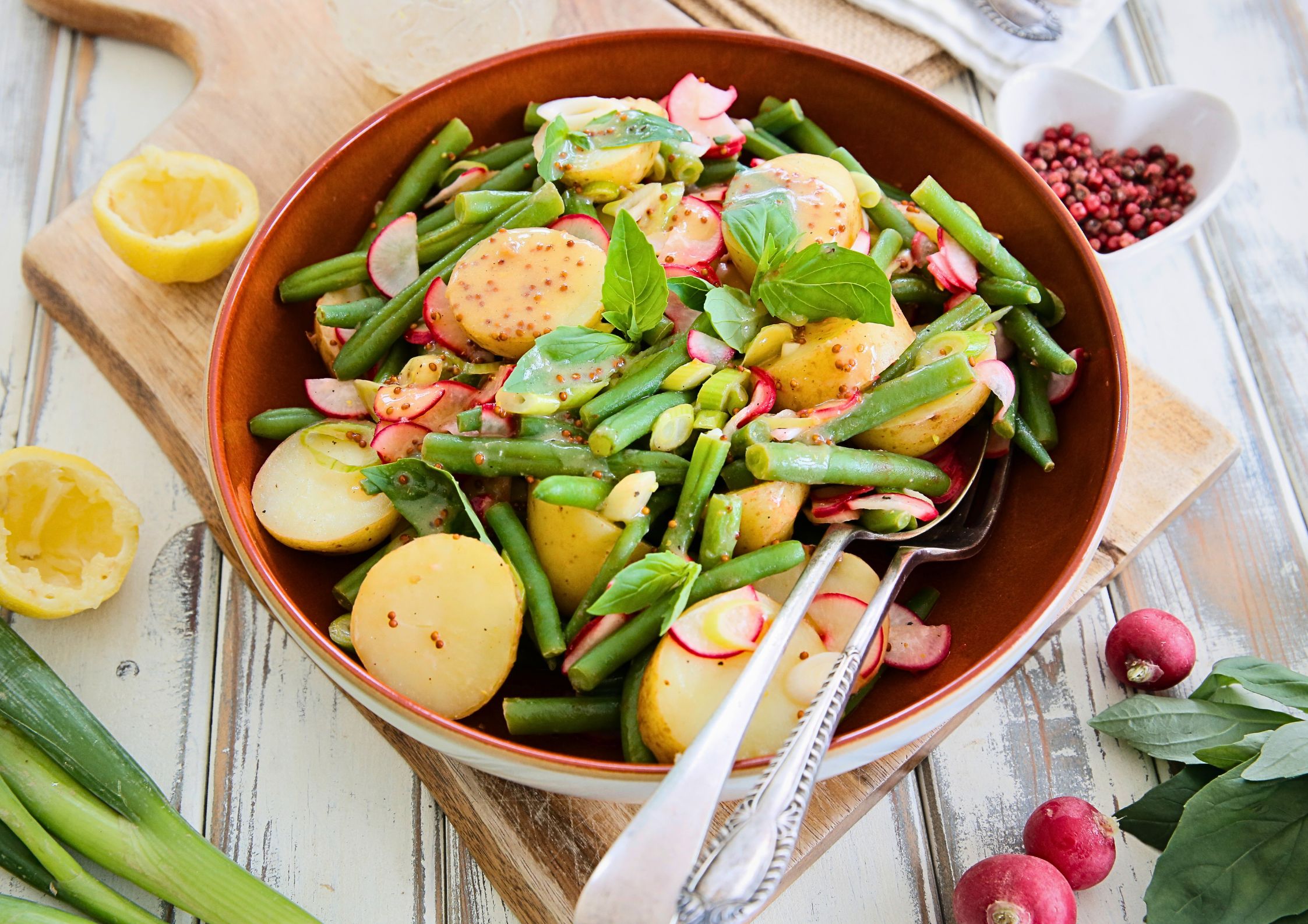 Warm spring salad in a brown bowl featuring boiled new potatoes, green beans, sliced radishes, and fresh basil, drizzled with a miso lemon dressing. Surrounded by fresh ingredients like radishes, spring onions, and lemon on a rustic wooden surface.